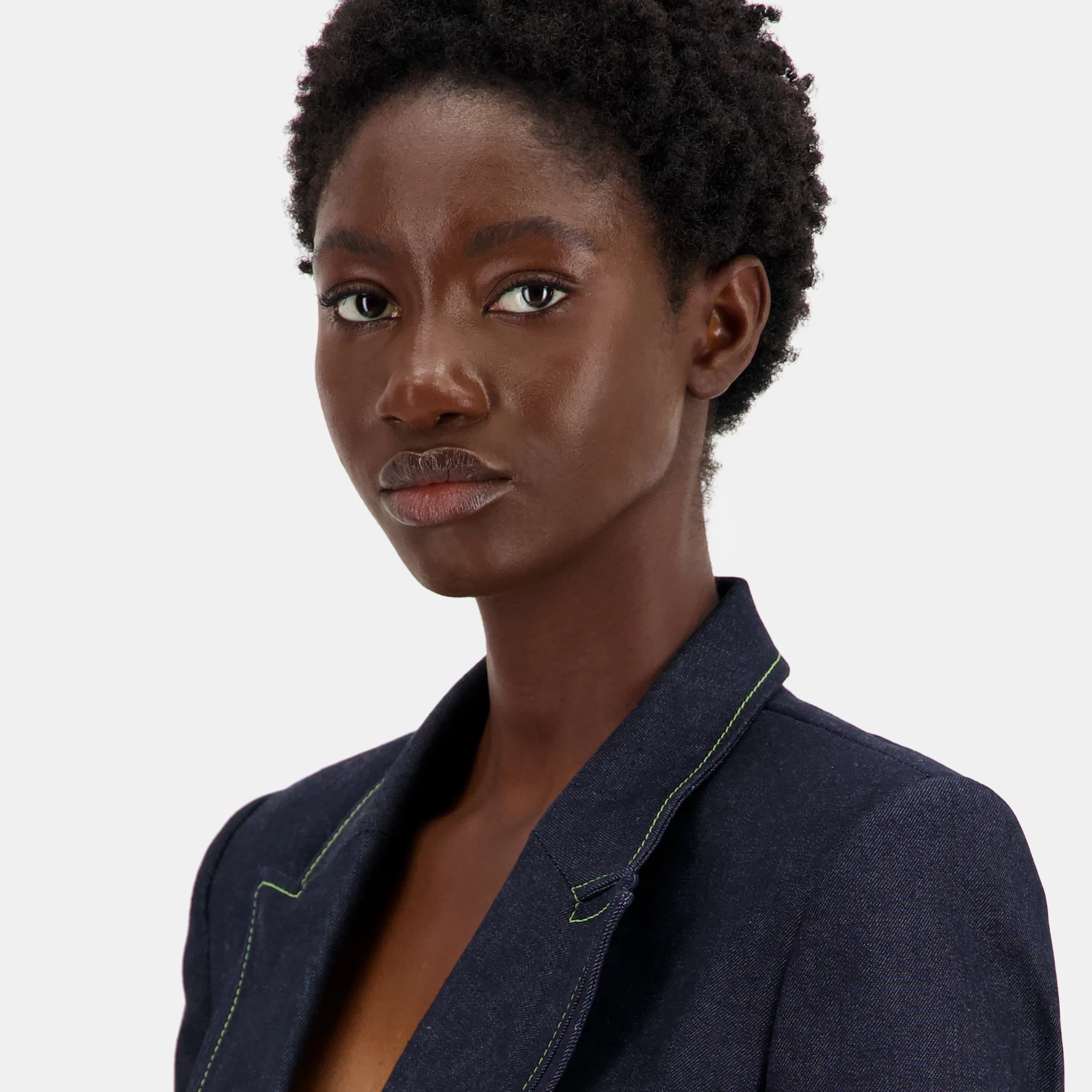 Model wearing a dark blue cropped blazer with powerful shoulders, contrasting light green pistachio stitching details, and black and white mother-of-pearl button fastening. Tailored from cotton denim with a slim fit and silk lining.